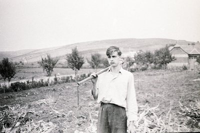A young man stands in a field, carrying a wooden pole over his shoulder. He wears a short-sleeved shirt and trousers, typical...