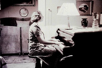 A woman in a patterned dress sits at an upright piano, playing. Interior scene shows a formal living room with dark wood furn...