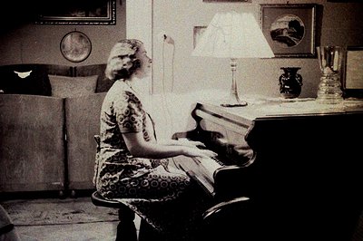 A woman in a patterned dress sits at a piano in a room with decorative elements. The room includes a cabinet, a large vase, a...