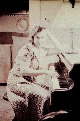 Vintage sepia-toned portrait of a woman playing a grand piano in an indoor setting, likely mid-20th century. She wears a patt...