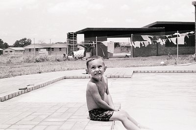 Black-and-white shot of a young boy sitting on a concrete pool ledge, smiling directly at the camera. Mid-20th century reside...
