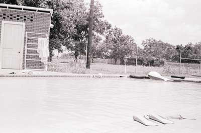 Empty concrete pool with discarded inflatable floats, likely mid-20th century. Brick utility building with white-trimmed door...