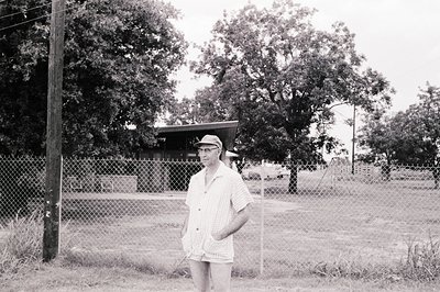 Mid-century street portrait: Man in 1960s-style cap and button-up shirt stands near chain-link fence. Background shows small ...