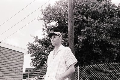 Mid-century man in a flat cap and checkered shirt stands outdoors beside a chain-link fence. Brick building and lush foliage ...