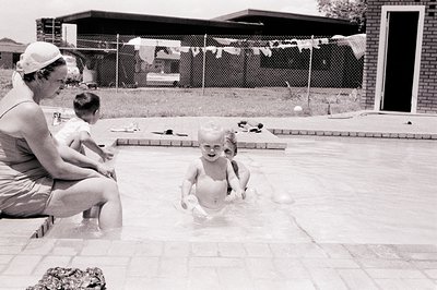 A mid-20th century backyard scene: a woman in a swimsuit and hat assists a toddler in a shallow pool, while another child pla...