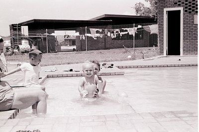Mid-century backyard scene with concrete pool, featuring three children in swimwear. Adult partially visible on left, seated ...