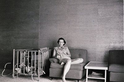 Mid-century modern living room with minimalist furniture. Woman seated on a tufted armchair beside a metal-framed playpen, ho...