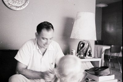 Mid-20th century black-and-white photo of a man in a white shirt working at a desk with a child seated in front. Decor includ...