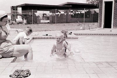 Mid-20th century backyard scene: three children playing in a shallow pool, one splashing water. Adult in white hat and dress ...