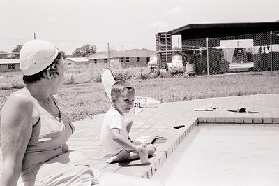 Mid-20th century poolside scene: woman in wide-brimmed hat and one-piece swimsuit supervises young boy holding a paddle. Conc...