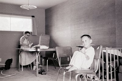 Mid-20th century classroom scene: young boy in rolled-up sleeves sits at a metal desk, smiling at camera. Adult at desk in ba...
