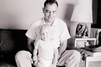 Mid-20th century black-and-white portrait of a man in a short-sleeve dress shirt and trousers holding an infant indoors. Mini...