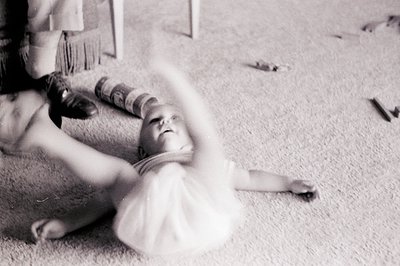 A candid black-and-white shot of an infant playing on a shaggy carpet, arms extended. Adult hand gently guides a rolled-up bl...