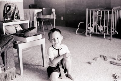 Mid-20th century indoor playroom with child seated on sand-filled floor. Wooden furniture includes a low table with books, a ...