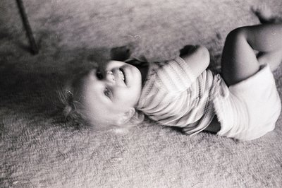 Black-and-white candid of a young child laughing while lying on a textured carpet, mid-20th century. Soft focus and warm ligh...