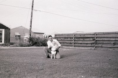 A man kneels on grass holding a young child in a mid-20th-century suburban yard, likely 1950s–1960s. Wooden fencing and utili...