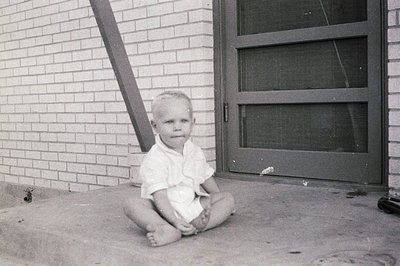Young child in mid-20th-century attire sits barefoot on concrete porch steps, dressed in a short-sleeve button-up shirt. Bric...