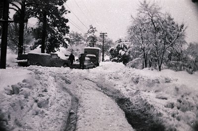 Mid-20th century black-and-white street scene buried under heavy snowfall. Classic vintage cars (1940s-50s) partially covered...