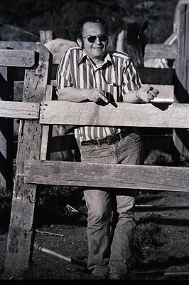 Mid-century man in retro striped shirt and sunglasses leans on wooden fence, likely 1960s–1970s. Rustic outdoor setting with ...