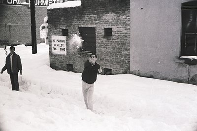 A man in mid-step uses a fire extinguisher on a brick wall in a snowy alleyway. "No Parking in Alley Fire Zone" sign visible....