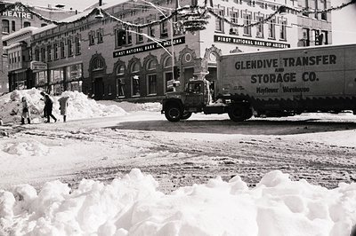 Mid-20th century urban winter scene with heavy snow accumulation. Prominent vintage truck labeled "Glendive Transfer & Storag...