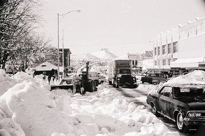 Mid-20th century winter street scene with heavy snow accumulation. Snowplows clear snow from a narrow road flanked by parked ...