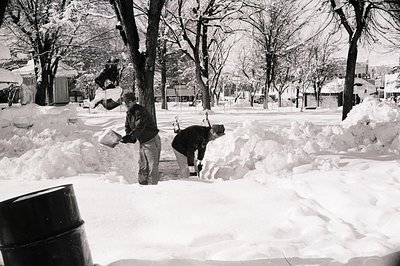 Two men clear snow in a mid-20th century urban park, using shovels and a wheelbarrow. Snow-covered trees and buildings sugges...