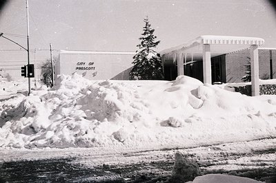 Mid-century municipal building in Prescot, buried under heavy snowfall. Brick facade with "City of Prescot" signage, flanked ...