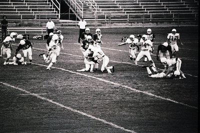 Vintage black-and-white football action shot: players in numbered jerseys (30, 31, 40) mid-play on a grass field with empty s...