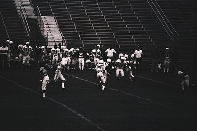 Black-and-white shot of an American football game in progress, featuring players in vintage uniforms (white helmets, numbered...