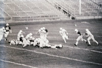Vintage black-and-white football action shot: players in helmets and jerseys clash mid-play on a grass field with empty stadi...