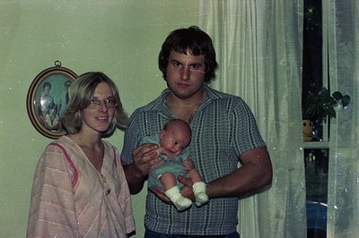 A 1970s indoor family portrait: a woman in a striped blouse and man in a checkered shirt pose with a baby in a light blue out...
