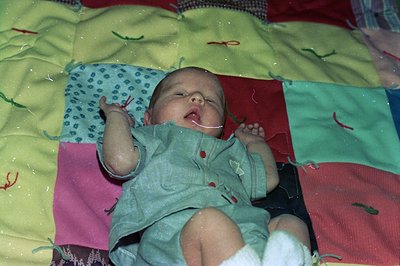 A newborn infant wrapped in a multi-colored quilt with geometric patterns, lying on a patterned blanket. The quilt features b...