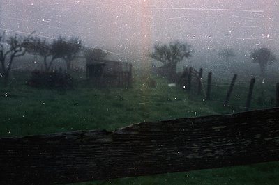 Rain-soaked rural landscape viewed through a window, showcasing a stone fence and blurred farm buildings in misty haze. The s...