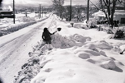 Black-and-white street scene showing a person clearing snow from a roadside ditch using a shovel. Snowbanks line both sides o...