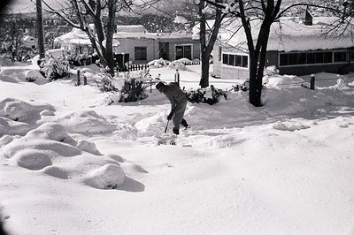 Mid-20th century suburban scene: man clearing snow with a shovel in a residential driveway. Snow-covered houses, bare trees, ...