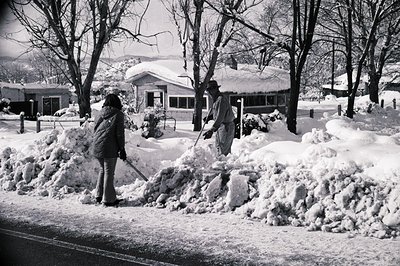 Two individuals shovel snow from a sidewalk piled high with drifts, surrounded by snow-laden trees and residential buildings....