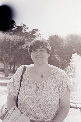 Black-and-white portrait of a woman in a floral blouse, standing outdoors near a fountain and trees. Mid-20th century fashion...