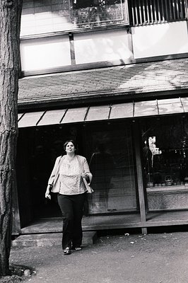 A woman in 1970s streetwear—blazer, patterned top, and wide-leg trousers—walks past a storefront with a metal awning and glas...