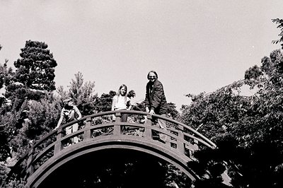 Three individuals pose on a curved wooden bridge in a lush, forested setting, likely a Japanese garden. The bridge’s arched d...