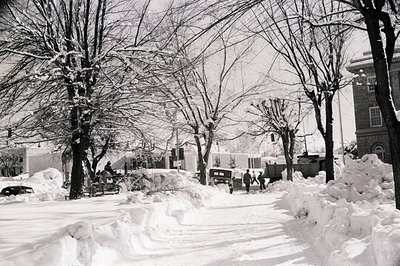 Snow-covered urban street with vintage cars, bare trees, and pedestrians in mid-20th century attire. Classic black-and-white ...