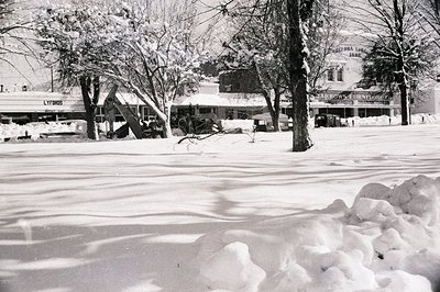 Mid-century American small-town street blanketed in snow, featuring "Lytods" and "Arrow Furniture" storefronts. Snow-covered ...