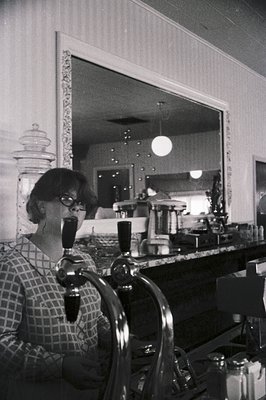 Mid-century bar scene with woman in patterned blouse sipping from a wine glass. Reflective mirror reveals vintage bar setup w...