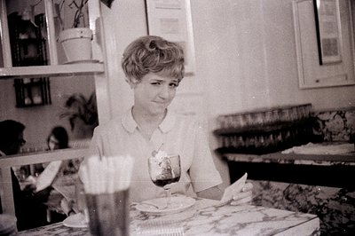 Vintage black-and-white photo of a woman seated at a table in what appears to be a mid-century café or home. She wears a shor...