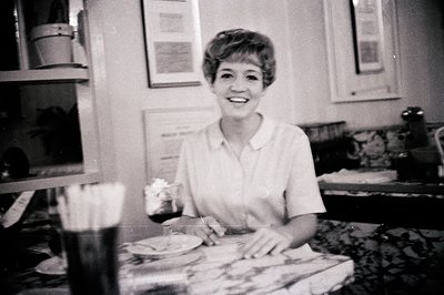 Mid-century kitchen scene featuring a woman in a short-sleeved blouse holding a knife, preparing food on a marble-topped coun...