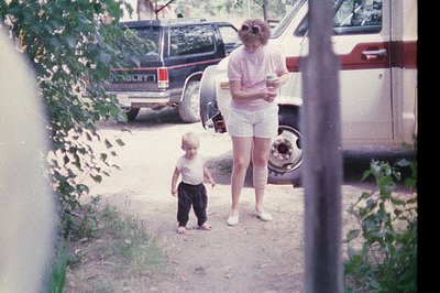 A woman in 1970s-style shorts and a blouse holds a child’s hand in a residential driveway. A vintage **Rover** truck with "RO...
