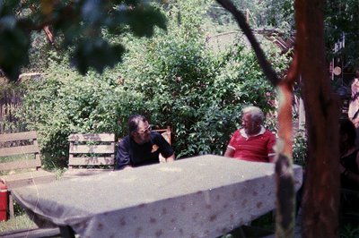 Two individuals seated at a covered picnic table in a lush, shaded garden, framed by foliage. The man in a dark shirt and wom...