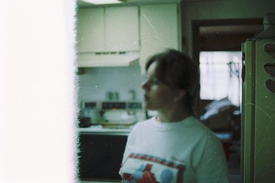 Vintage Polaroid-style shot of a person in a kitchen, blurred for privacy. White T-shirt with red/blue graphic, standing near...