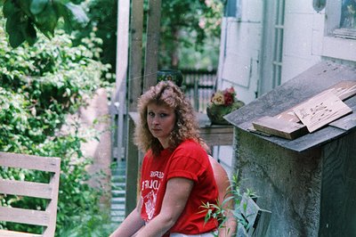 Vintage candid of a person in a red T-shirt (text partially visible: "FRUIT") leaning on a rustic wooden railing in a lush, o...