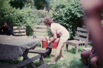 Vintage outdoor scene featuring mid-century picnic setup. A person in light-colored pants and a white top kneels beside a red...